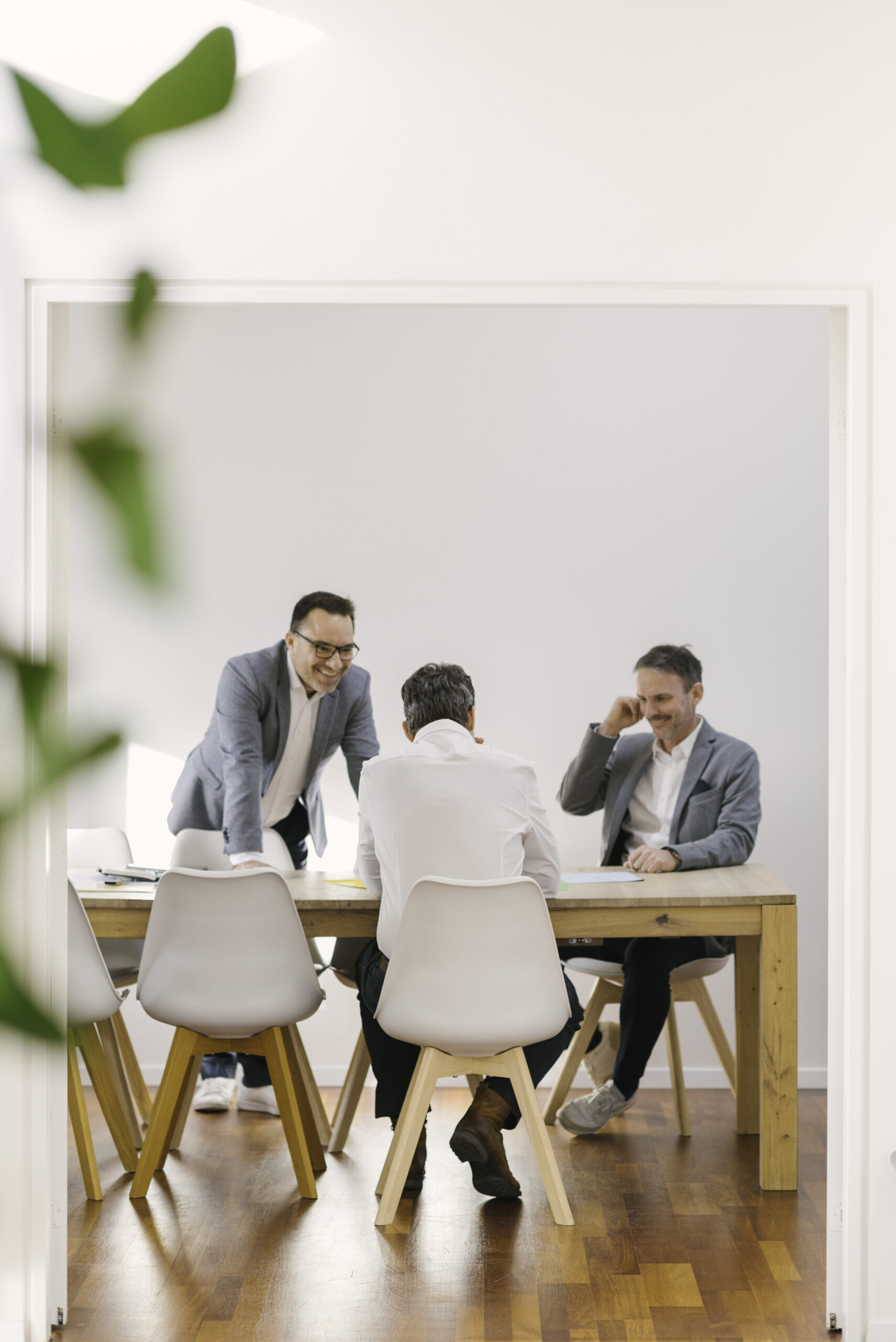 Bild von drei Männern im Sitzungszimmer. Einer steht und lacht. Die beiden anderen Sitzen am Sitzungstisch.